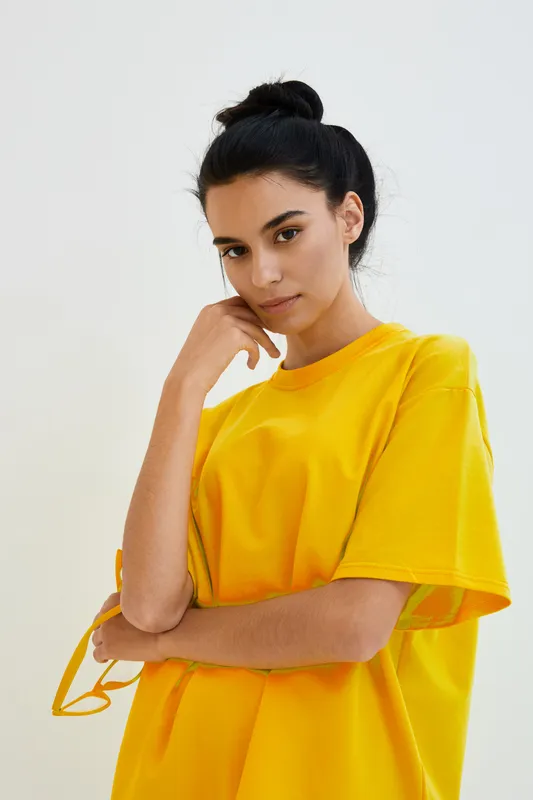 Young woman wearing a bright yellow oversized shirt with a thoughtful expression and casual hairstyle against a clean neutral background, showcasing modern minimalistic style.