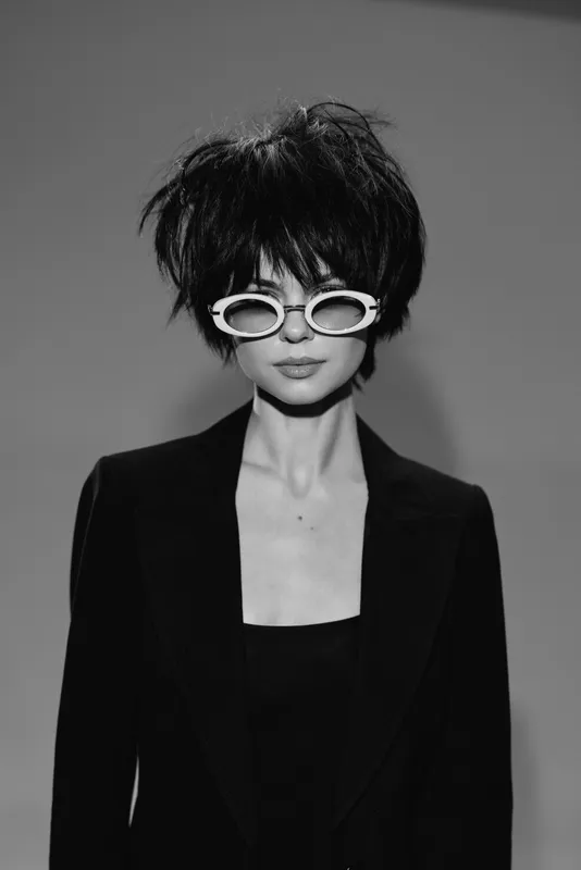 Black and white stylish woman with short hair wearing trendy sunglasses and dark blazer posing confidently in studio lighting.