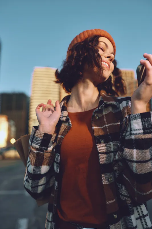 Woman portrait in urban street at golden hour, joyful and candid with smartphone, beanie and plaid jacket. Emotional storytelling, authenticity and mindful living vibe in modern city lifestyle.