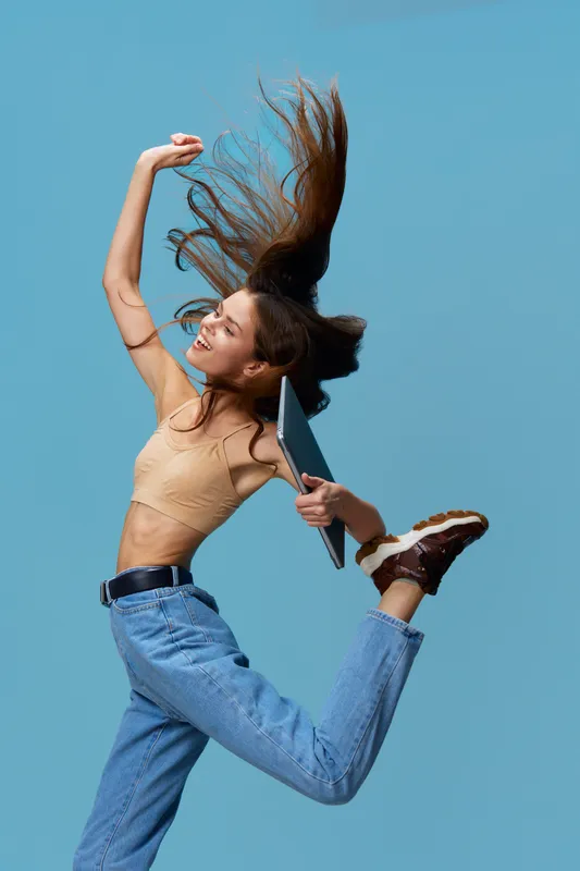 joyful young woman jumping with laptop in hand against blue background, wearing casual jeans and beige top, dynamic pose with flowing hair, studio lighting.
