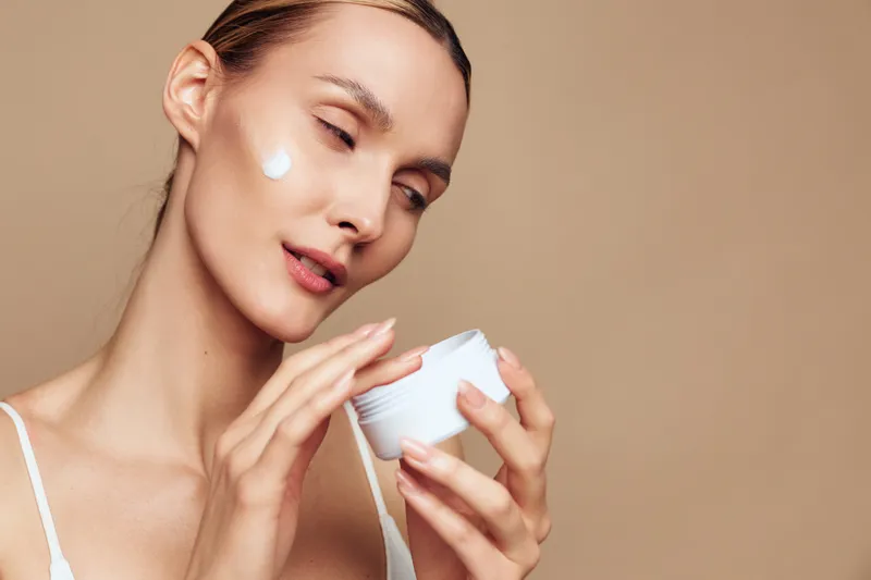 Young woman with brunette hair applies moisturizing cream on her cheek while holding a white jar against a beige background in a skincare routine moment.