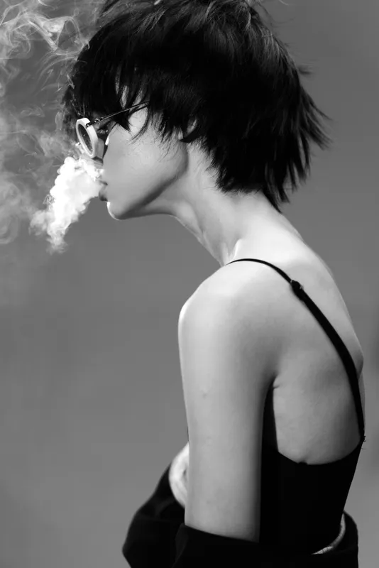 Black and white portrait of a young woman exhaling smoke, featuring short hair, glasses, and a stylish outfit with a dark background.