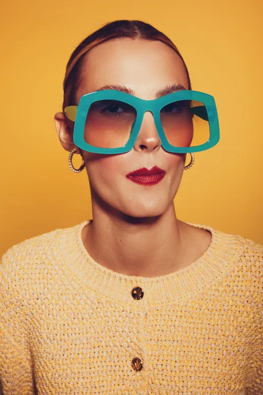 Stylish woman wearing large blue sunglasses and yellow sweater poses against orange background. Fashion portrait with bold accessories and confident expression in studio lighting.
