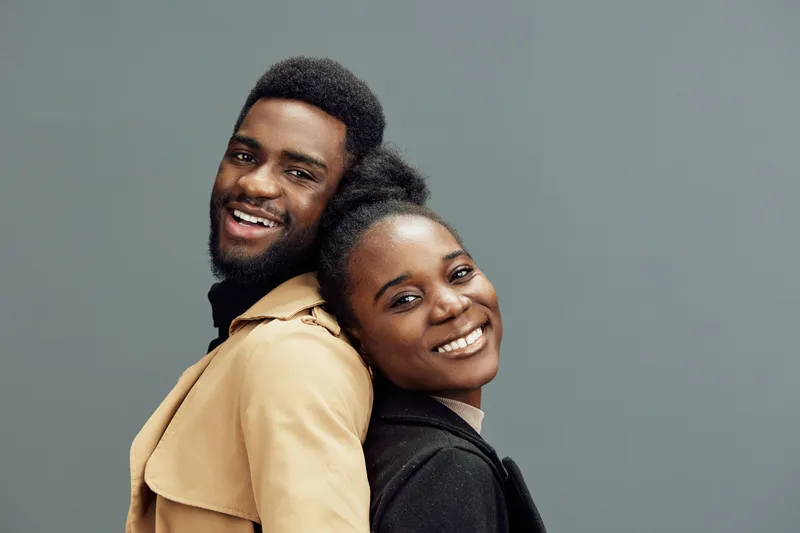 Smiling young African couple showcasing modern fashion against a neutral gray background, exuding joy and confidence in their vibrant outfits