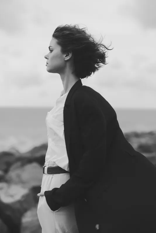 Woman standing in a windy coastal scene, wearing a coat and white outfit, facing the horizon, in a monochrome mood with a calm sea behind and a sense of distant horizon