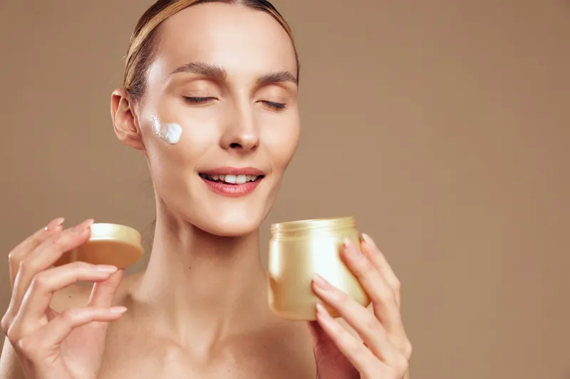 Young woman with blonde hair smiles softly while applying white cream to her cheek, holding two jars in a soft beige studio background, conveying a calm skincare moment.