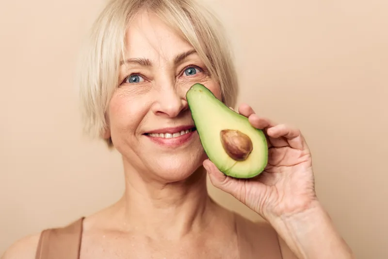 Smiling elderly woman with short blonde hair holds half an avocado close to her face on beige background. Concept of healthy eating, natural beauty, wellness, and positive lifestyle.