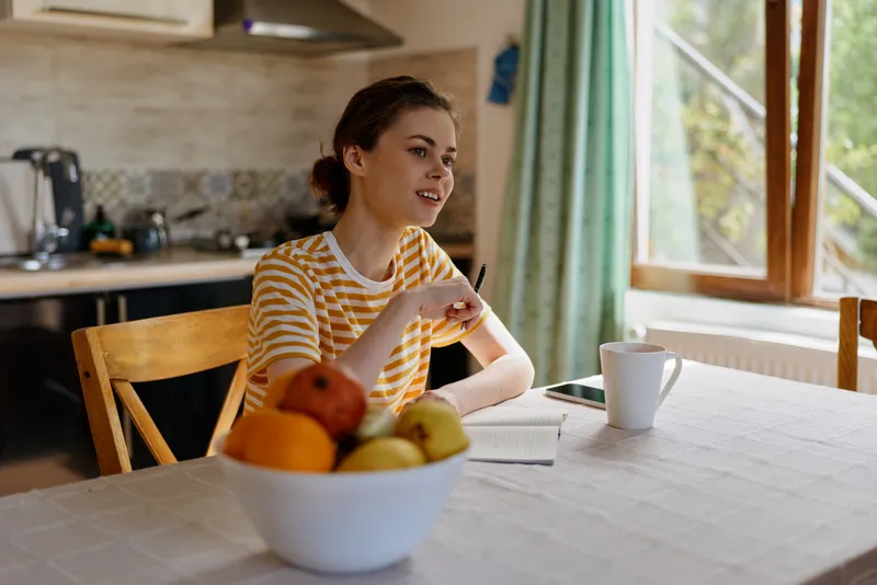 Cheerful woman at a bright kitchen table, wearing a striped top, speaking softly, sunlight flooding the space, fruit bowl in foreground, warm relaxed home lifestyle moment