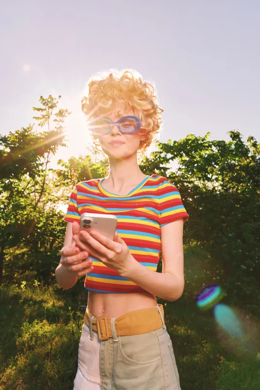 Cheerful woman lifestyle outdoors retro color sunglasses striped casual summer sunlight, scrolling on a phone in a sunny moment, relaxed pose, lush greenery in the background