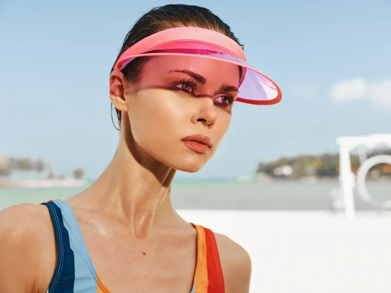Portrait of a focused female athlete at the beach wearing a color block swimsuit and a pink visor, sunlit skin and calm blue water in the background.