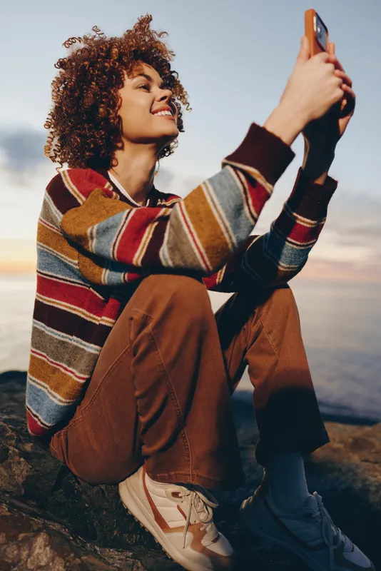 Smiling woman lifestyle outdoors in winter, striped sweater, holding smartphone for a selfie, travel on rocky mountains trail, boots and backpack, sunset light