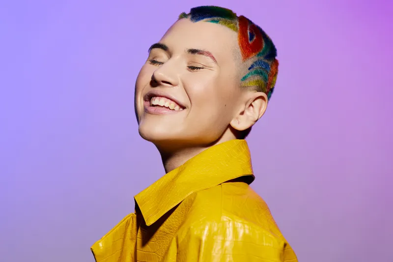 cheerful woman with colorful short hair and vibrant yellow jacket smiling with eyes closed against purple background, expressing happiness and creativity