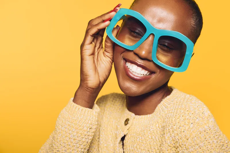 Happy smiling black African woman with dark skin wearing oversized blue glasses and beige sweater poses on yellow background. Concept of joy, fashion, and positive mood with vibrant colors.