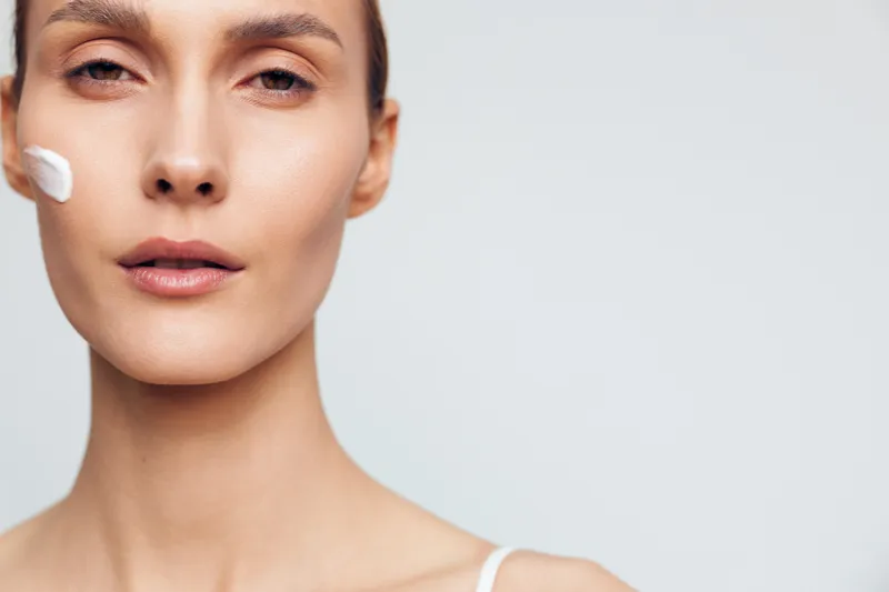 Woman skincare beauty portrait, face cream on cheek, cosmetic studio treatment, fresh smooth skin concept, close up calm expression, soft lighting, neutral gray background, clean look