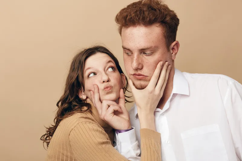 Young woman playfully pinches the cheeks of a serious young man against beige background. Concept of fun, relationship, friendship, and lighthearted moments between couple.