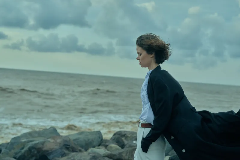Windy coastal scene of a woman on rocks by the sea, coat billowing behind, calm expression, looking toward the horizon under moody clouds, outdoor fashion mood with natural elements.