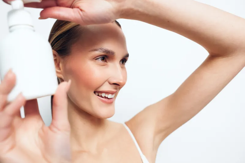 Young woman with clear skin smiles while applying moisturizer to her face in a bright skincare routine at home with a white background.