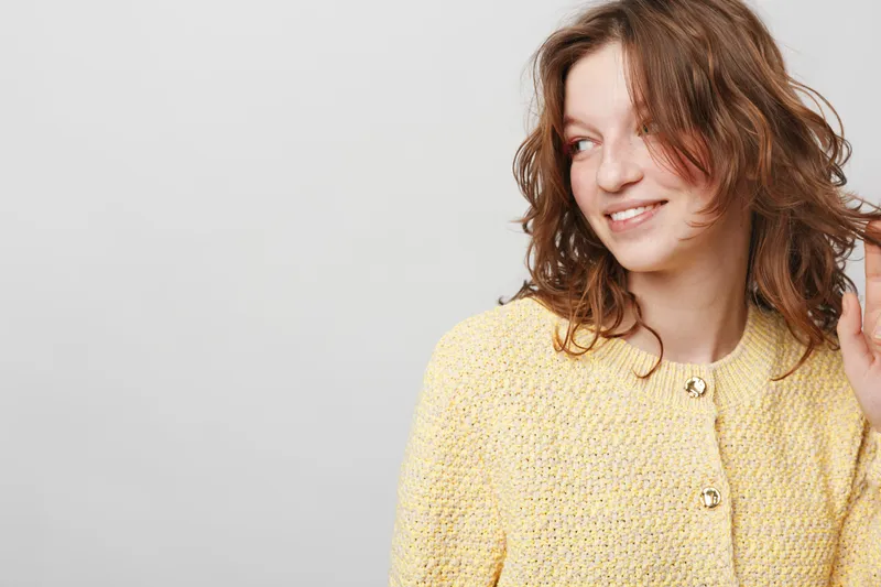 Smiling young woman with curly brown hair wearing yellow textured jacket looks away on grey background. Concept of casual style, happiness, and natural beauty portrait.