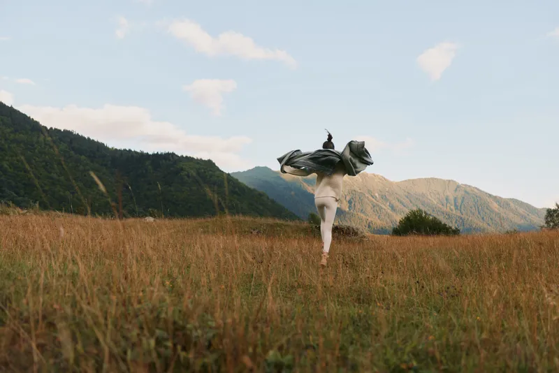 Woman lifestyle outdoors running through tall grass field with flowing jacket mountains background clear sky natural light freedom and movement