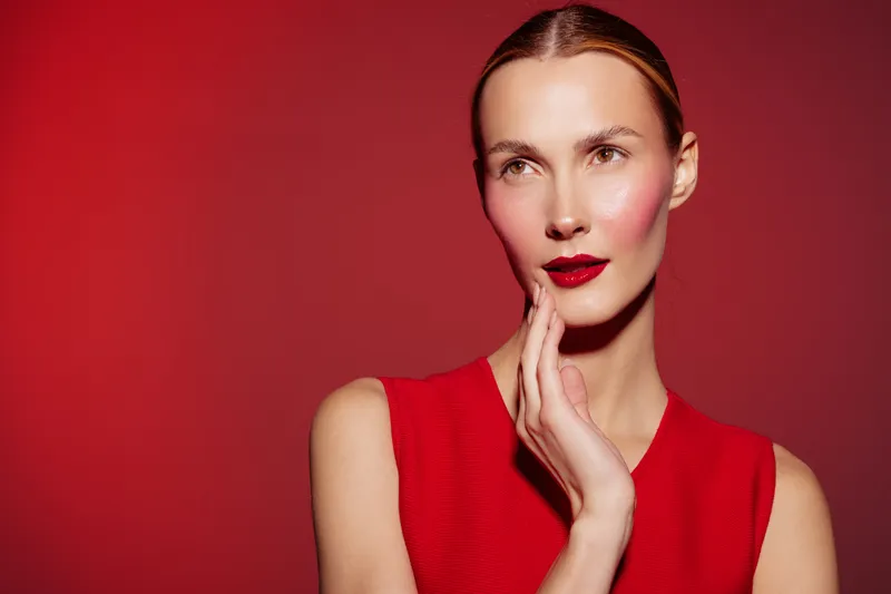 Portrait of a thoughtful woman in red dress with red lipstick and blush on red background. Concept of fashion, beauty, makeup, and elegance in studio lighting.