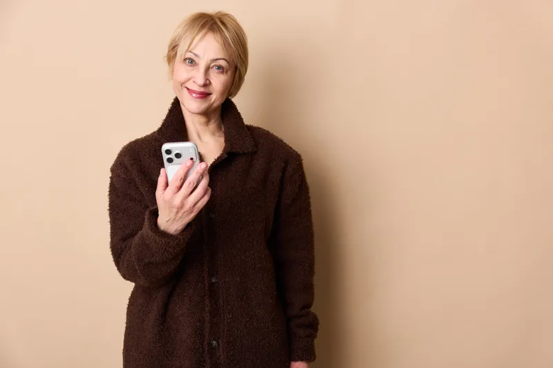 Elderly woman smiling, holding smartphone in hand, relaxed portrait against beige background, showing communication and technology adoption, social media concept, social connection