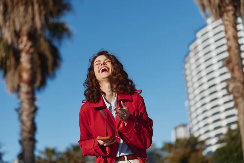 Young woman with curly brunette hair in a red jacket laughs outdoors in a bright urban setting with palm trees and tall buildings, capturing a carefree sunlit moment.