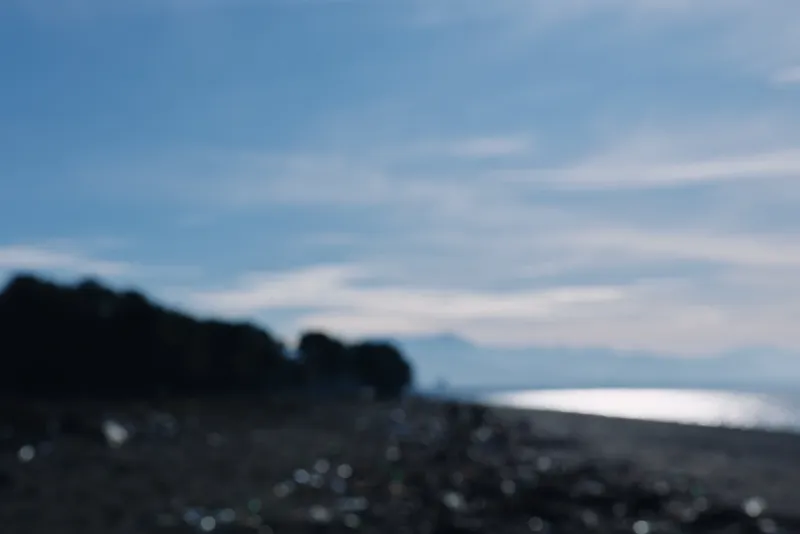 Blurred beach scene with calm ocean and distant horizon under blue sky, pebbled shore and soft silhouette trees, creating a tranquil coastal mood and sense of quiet reflection.
