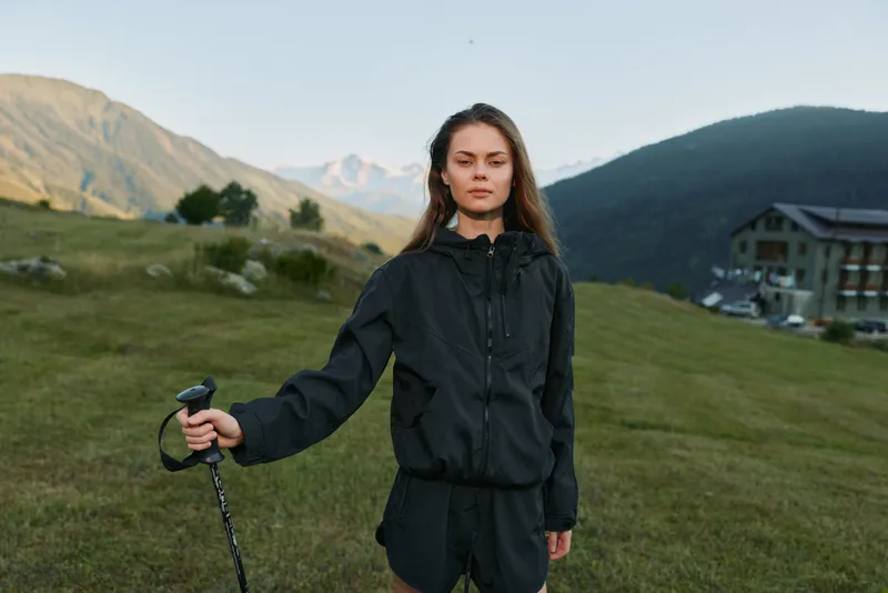 A focused woman stands outdoors in a rugged countryside, holding a trekking pole. She wears a dark jacket and shorts, gazing ahead with confidence in a wide, open landscape.
