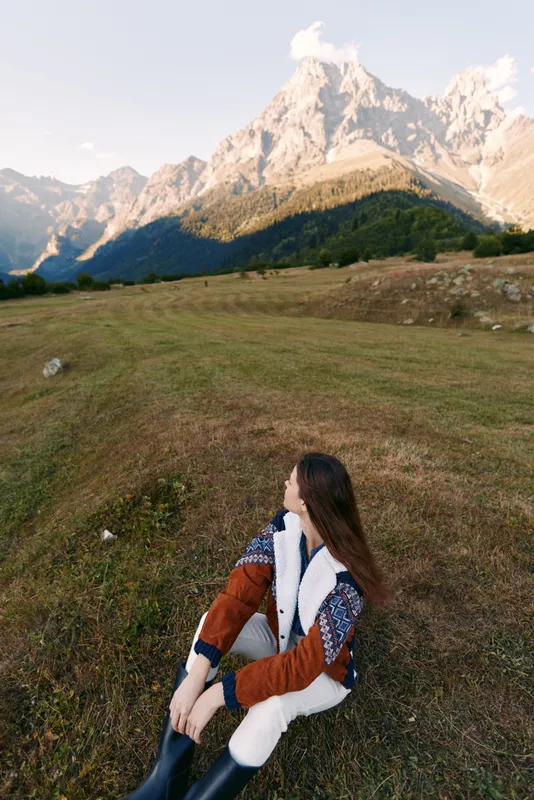 Woman in autumn sweater sits in a mountain meadow, nature landscape with high peaks and soft light, travel and hiking scene, peaceful outdoor solitude and scenic horizon.