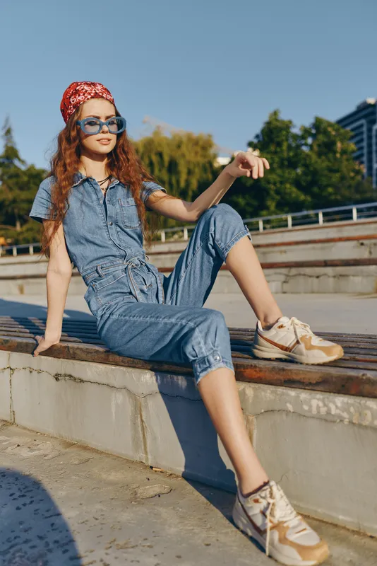 Lifestyle woman in modern boho-western denim street style sitting outdoors with sunglasses and a red bandana in warm film color tones, relaxed urban fashion vibe.