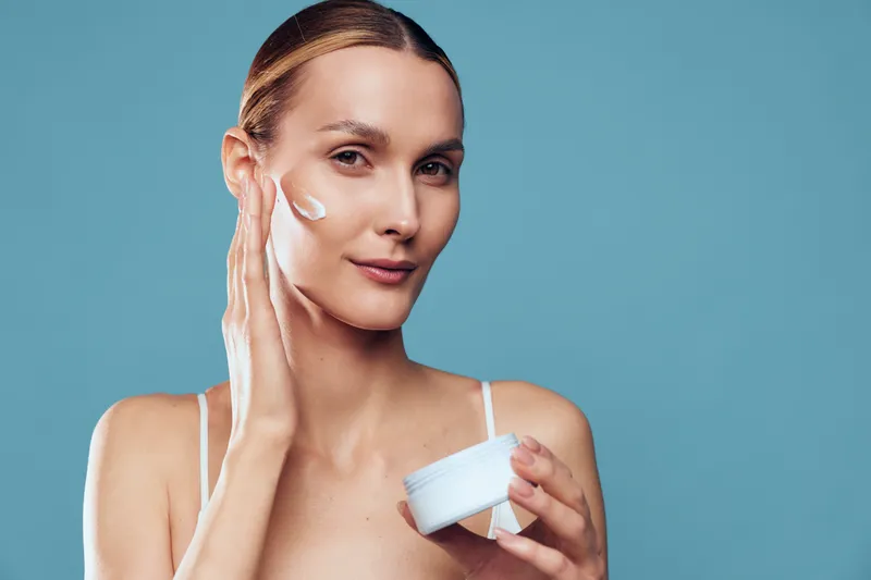 A young woman with clear skin applies moisturizing cream to her face in a close-up portrait against a blue background. Concept for skincare, beauty, and self-care routine.