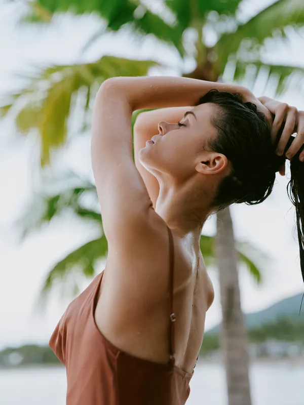 Sunlit portrait of a woman in a tropical setting, arms raised, hair swept back, wearing a warm brown swimsuit, smooth skin, and palm trees blurred in the background.