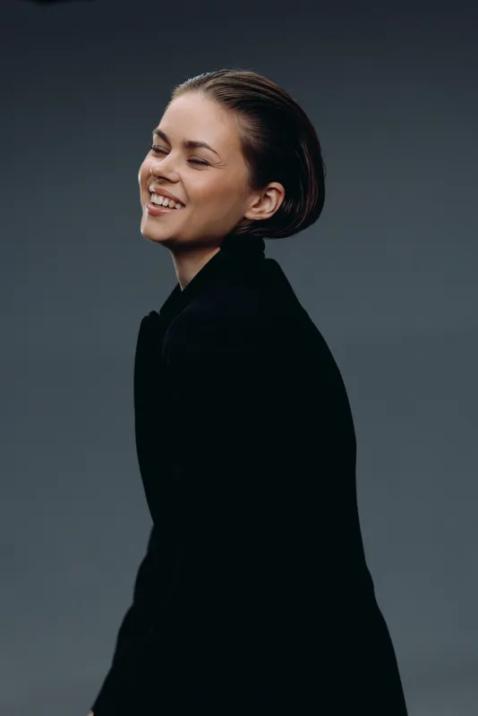 Smiling woman in black jacket with short hair expressing happiness and confidence on neutral dark background.