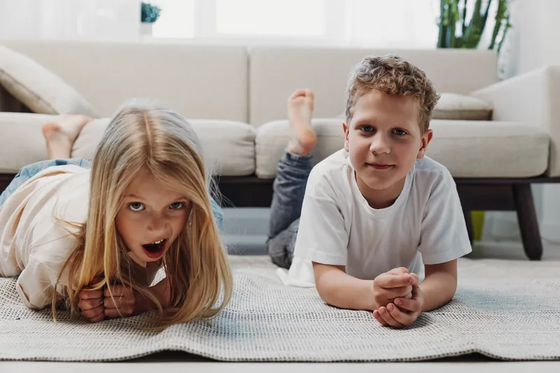 Two children lying on carpet in modern living room, smiling and relaxed. Casual home environment with natural light, cozy sofa, and indoor plants in background.