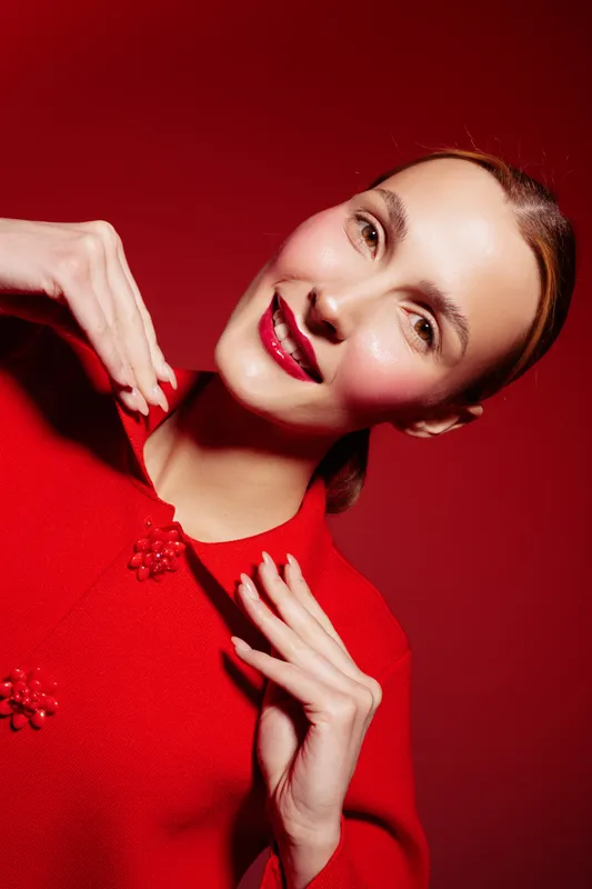 Smiling woman in red sweater poses against red background, showing joyful expression and bright lipstick. Concept of fashion, beauty, and vibrant style with warm lighting.
