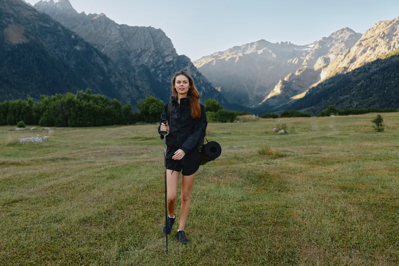 A young woman hikes across a wide alpine meadow amid towering peaks, using a trekking pole. She wears practical gear and carries a backpack, exuding focus and outdoor enthusiasm.