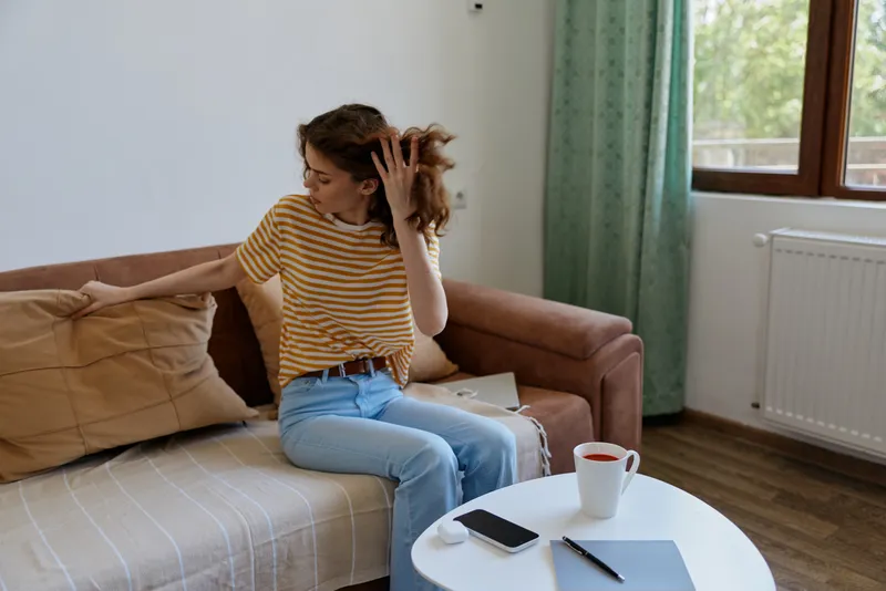 Casual woman sitting on a sofa at home, adjusting her hair in a relaxed mood. Natural light from a window, cozy living room, simple decor, everyday self care and comfort.