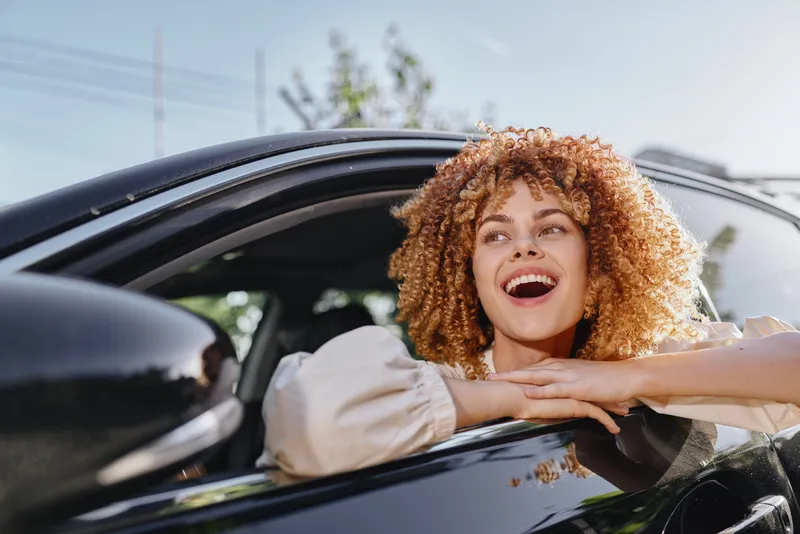 Smiling woman with curly hair enjoying a sunny day leaning out of the car window, expressing happiness and freedom in natural light outdoors.