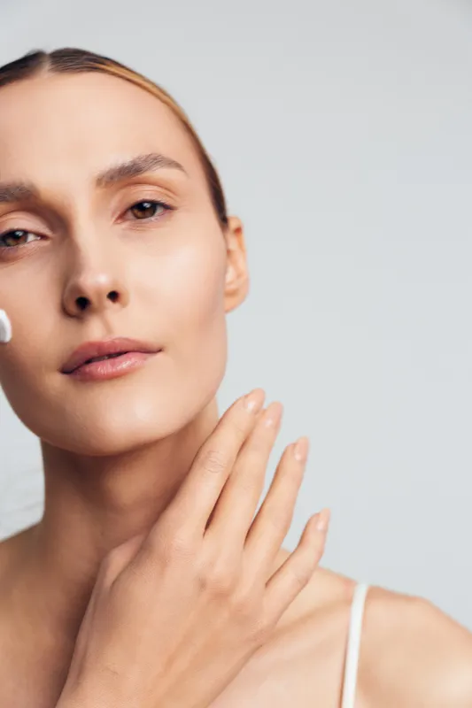 Young woman with clear skin applies white face cream on her cheek while gently touching her neck against a light background in a skincare routine moment.