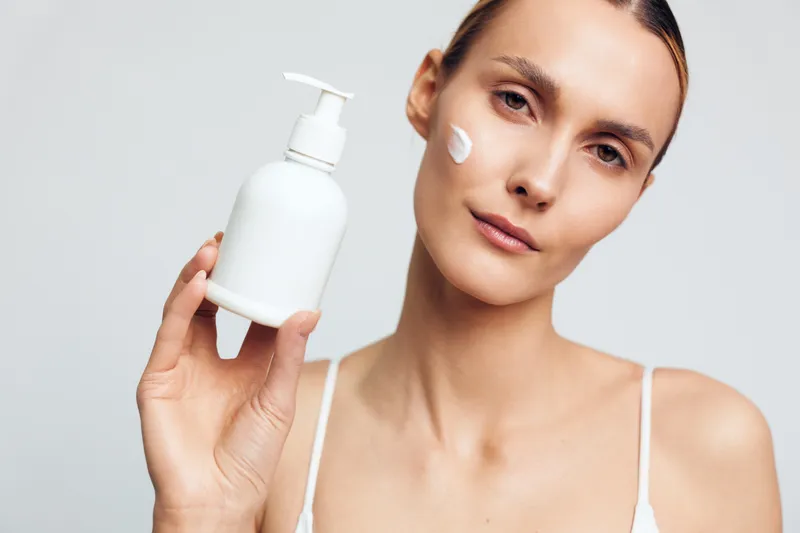 Young woman holding white bottle with pump dispenser applying cream on face closeup skincare routine natural beauty clean skin minimal background
