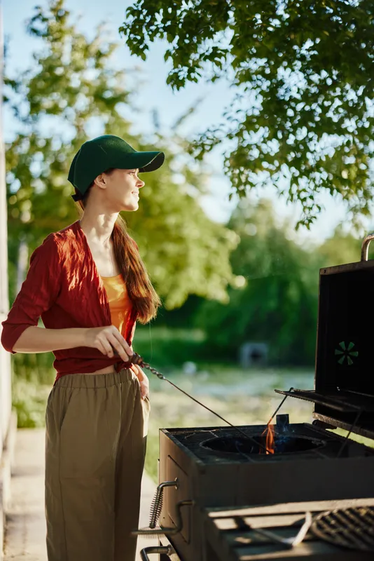 A young woman grilling outdoors, wearing casual clothing and a baseball cap, capturing the essence of summer and enjoyment in nature
