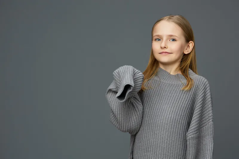 Charming young girl in oversized gray sweater, confidently smiling against a minimalist gray background, perfect for seasonal fashion and children s wear concepts