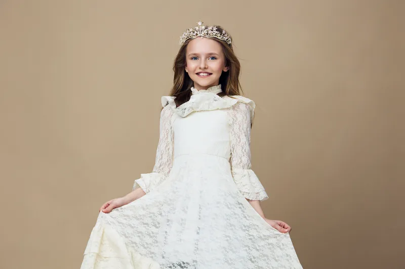 Smiling girl in a white lace dress and crown against a beige background, exuding joy and elegance, perfect for children s fashion and royal themed projects
