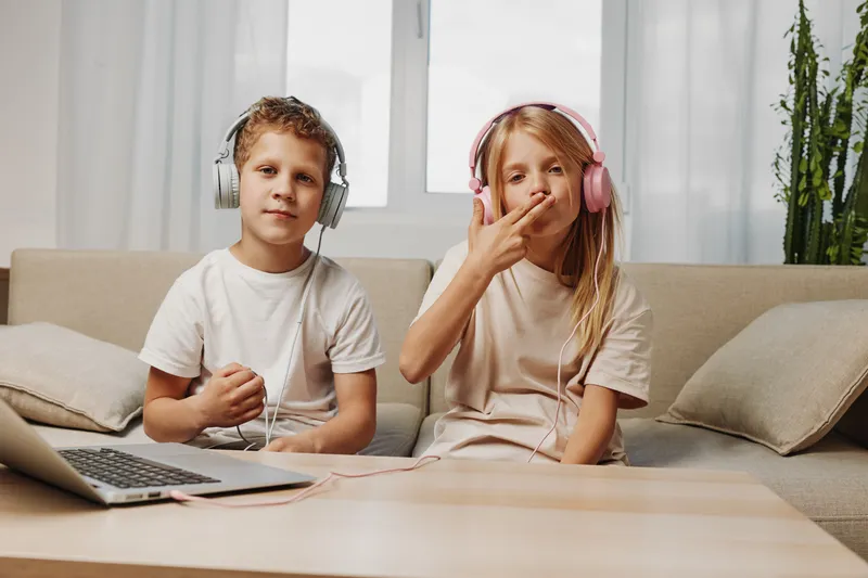 Two kids sitting on a couch wearing headphones, enjoying music together with a laptop on the table. Casual home environment with natural light through window.