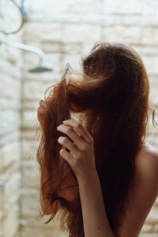 woman brushing hair under shower with stone wall background, natural light highlighting wet strands and peaceful morning routine moment