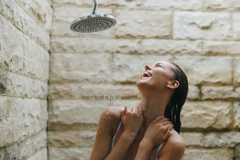 young woman enjoying outdoor shower with closed eyes and water droplets, relaxing under stone wall, natural light enhancing peaceful summer moment