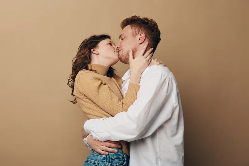 Young couple in casual clothes embraces and kisses tenderly on beige background. Intimate moment of love, affection, and connection between man and woman in warm studio lighting.