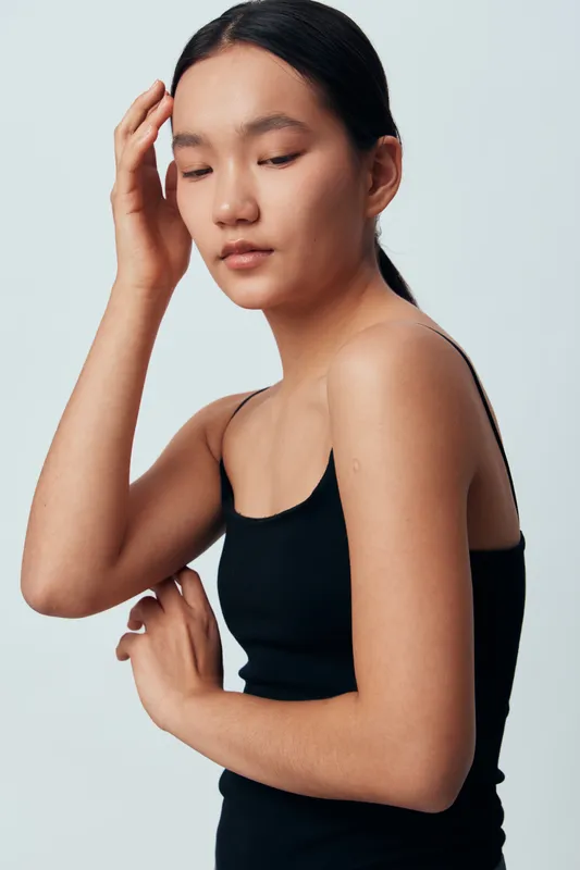 Young woman with closed eyes and calm expression touching her head and arm in black sleeveless top, posing against plain light background for beauty and wellness concept.