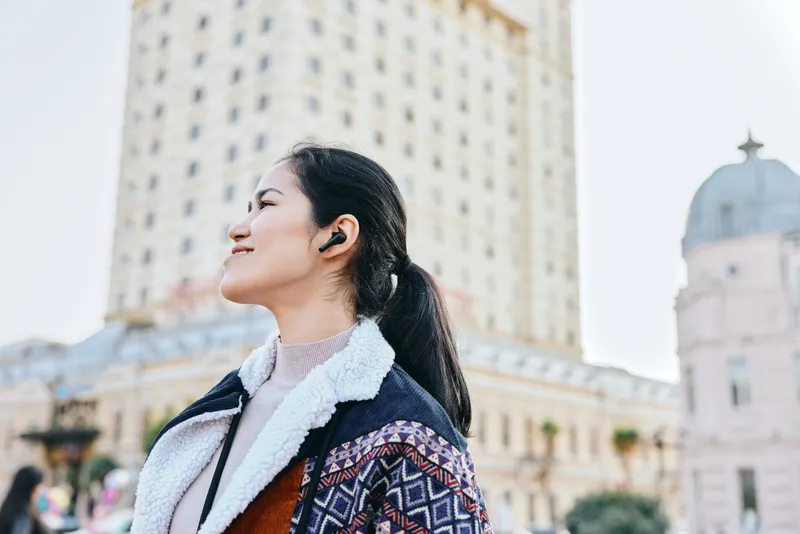 Woman in stylish coat with shearling collar gazes upward in an outdoor city setting, wearing headphones, with modern architecture and bright sky creating a candid urban lifestyle moment