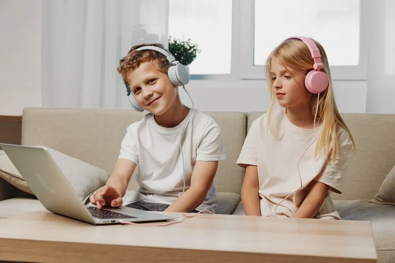 Two children with headphones use laptop at home sitting on couch. Concept of remote learning, entertainment, and digital technology for kids in casual room setting.